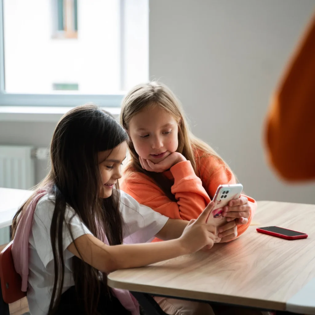 Two young girls are seated at a table, looking at a smartphone