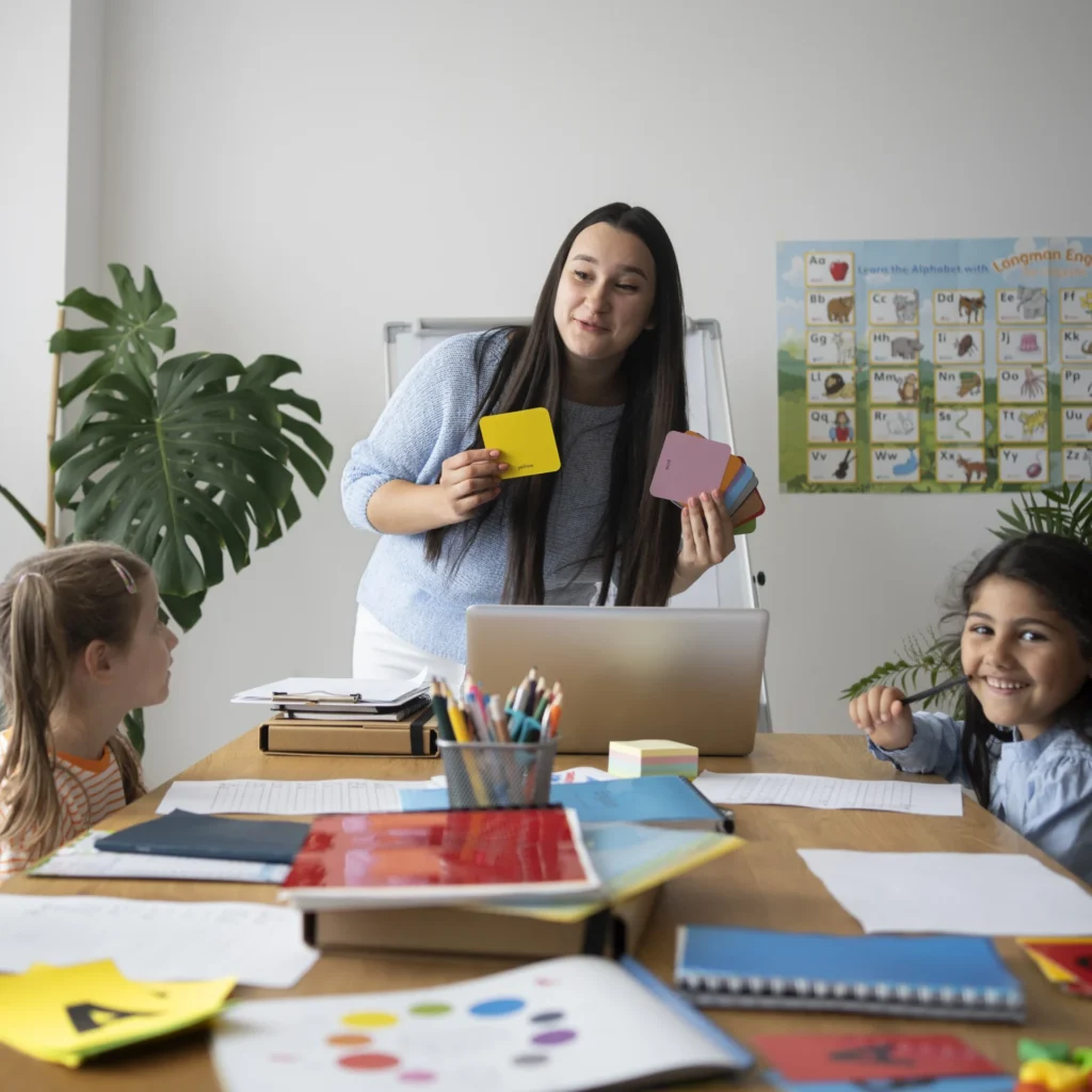 A classroom scene depicts a teacher presenting colorful shapes to a group of children seated around a table.