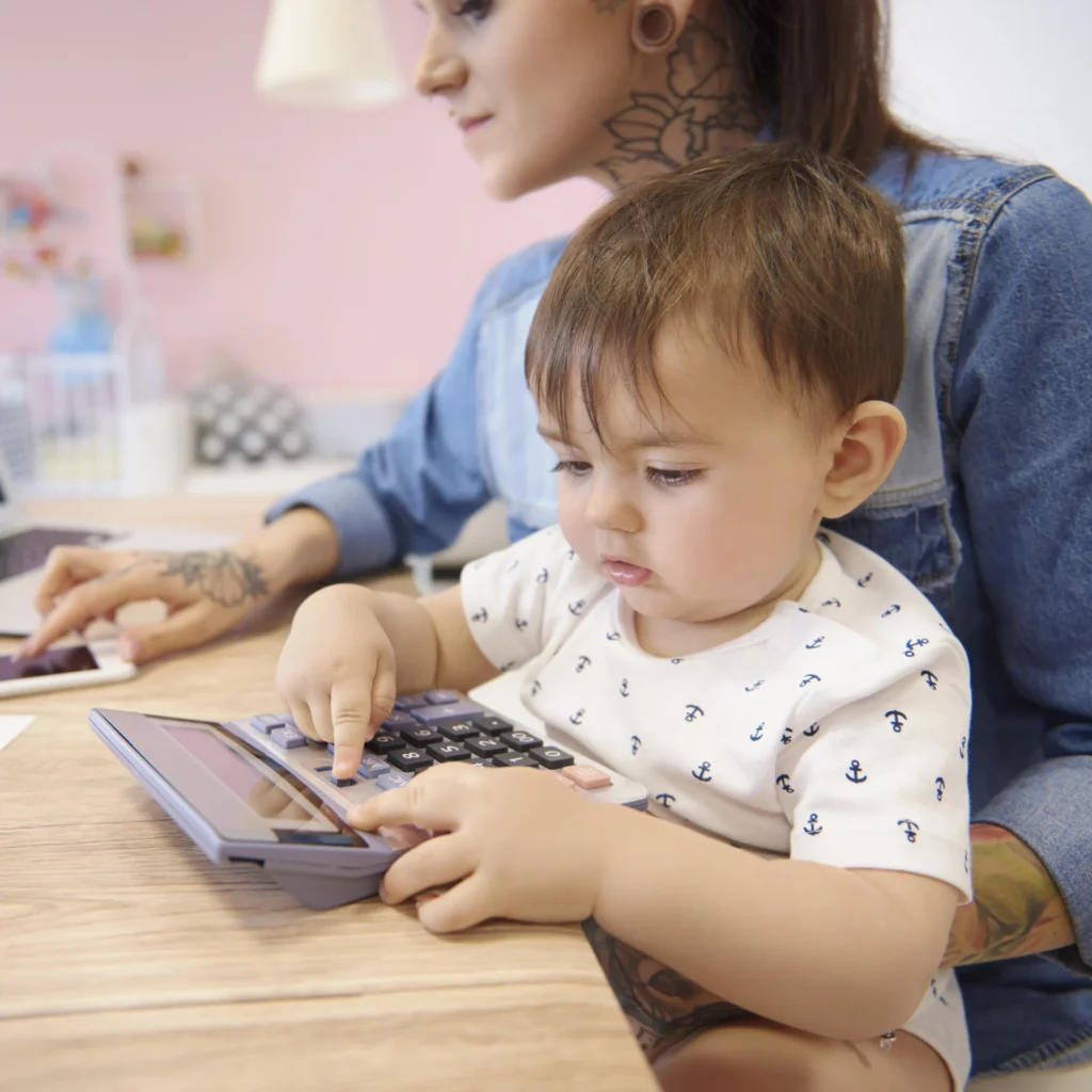 A woman sits at a table with a baby in her lap, both interacting with a calculator.