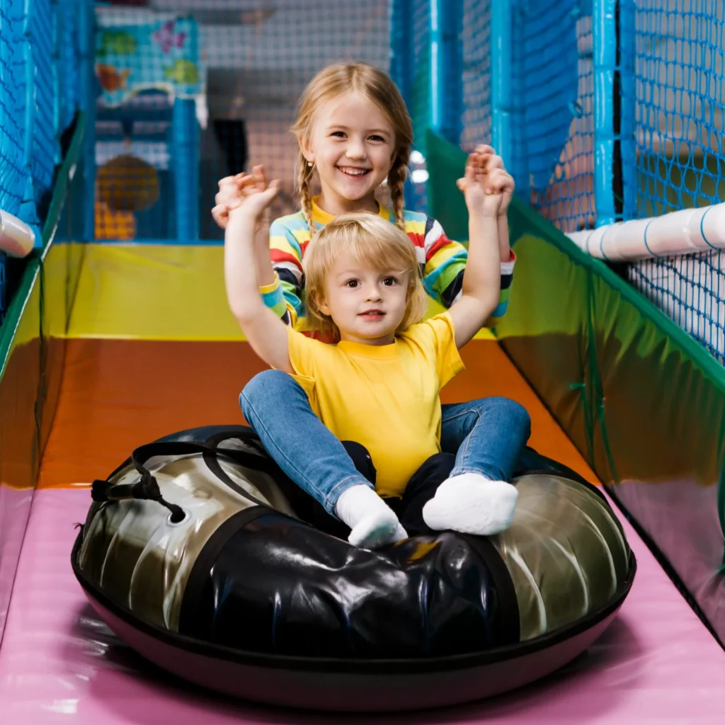 Two children smiling while sliding down on an inflatable tube inside a colorful indoor play area